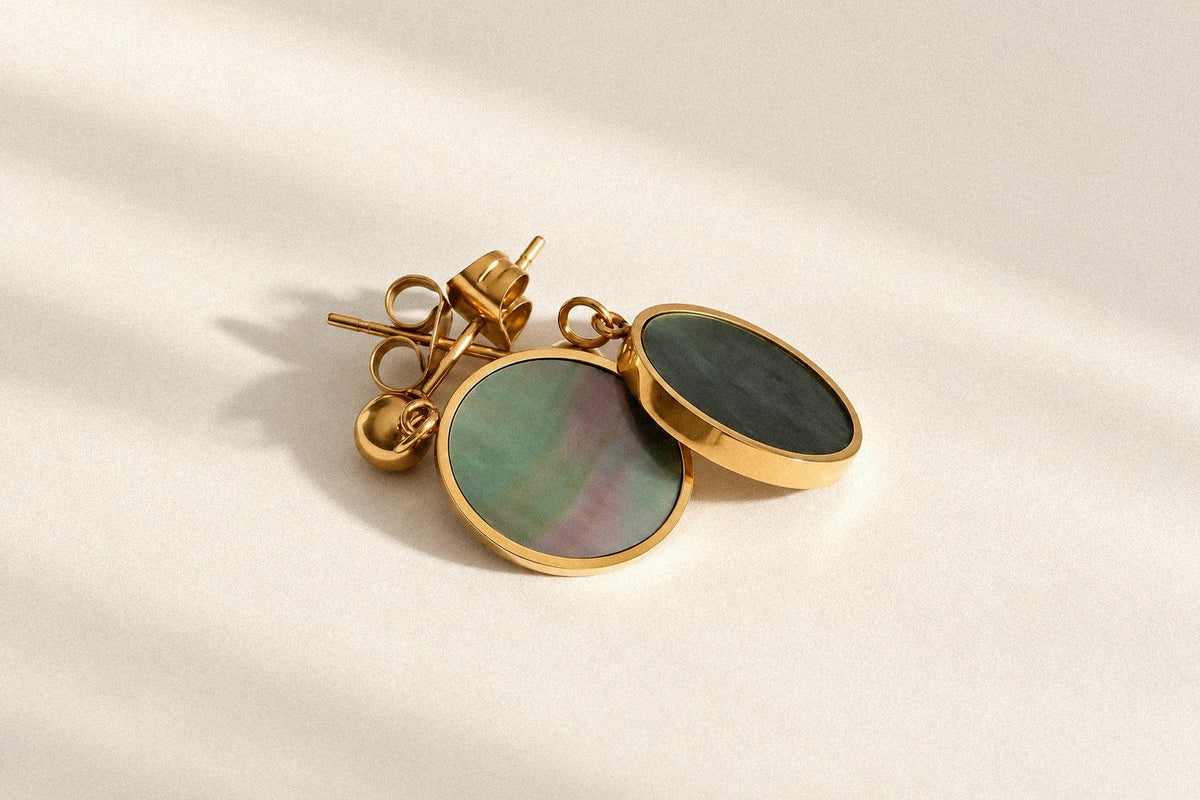 Mist Tide Stud Earrings with gold-colored rims and iridescent shell discs on a warm ivory surface.