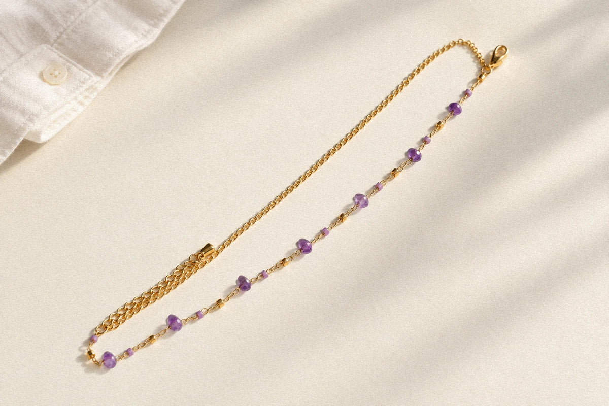 Purple gemstone bracelet laid diagonally on an ivory tabletop beside a cream shirt cuff.