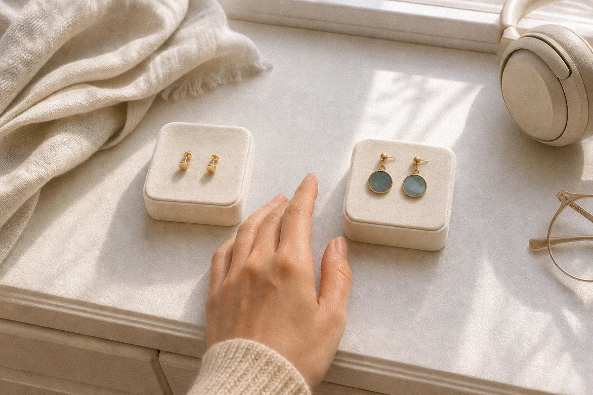 A hand choosing between gold studs and round mother-of-pearl drop earrings on ivory jewelry pads