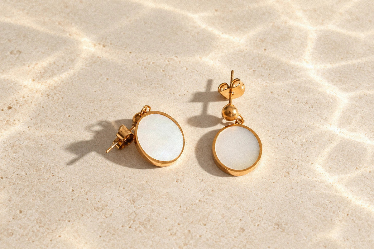 Tide Stone earrings with white mother-of-pearl inlays and gold-colored frames on an ivory stone surface with soft ripple-like light.